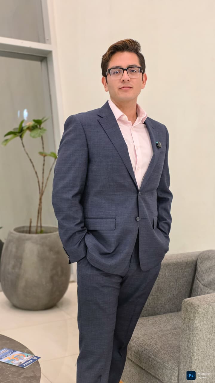 Young professional wearing a navy suit and glasses, standing with hands in pockets in a modern office lobby next to a gray sofa and potted plant.